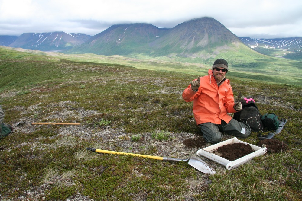 person kneeling in a tree-less plain near a shovel and hole in the ground; mountains in the distance