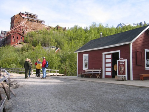 Group of people talking outside historic, red building, with large, red mill building and forest in background.