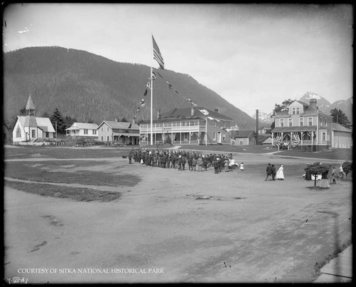 People gathered on the parade ground with Marine Barracks and Officers Sitka Town decorated with flags and bunting.