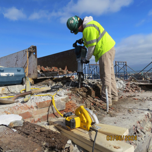 View of worker in high visibility gear removing bricks at the top of the Monument. Scaffolding is visible in the background and tools are staged in the foreground.