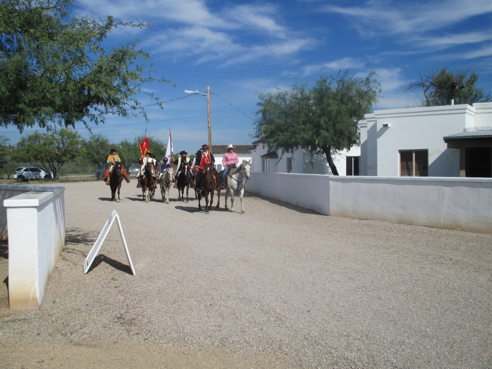 A group of people wearing Spanish colonial costume on horseback ride down a gravel road past a white stucco building