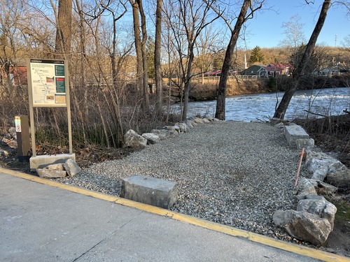 A gravel path lined with large rocks and trees leads to the river. To the left is an information panel. 