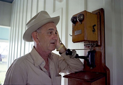 Wearing a Stetson hat, President Johnson speaks into an antique phone hanging on the wall.