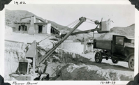 This is an historic black and white photograph from the Scotty's Castle Historic Photograph Collection, Death Valley National Park of large power shovel in excavated pit in the foreground ready to dump load into a dump truck parked on the road above. Spanish style structure in background to the left with a curved retaining wall running into the background. Inscriptions in black ink along upper and lower border.