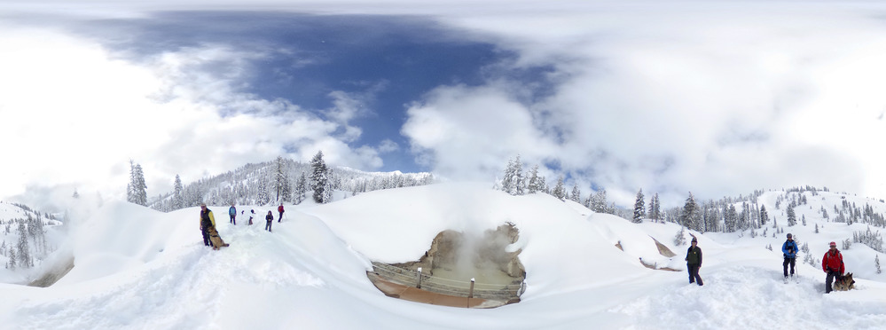 Eight people standing, spread out on snow with a steaming grey pool in the center in a panorama photo