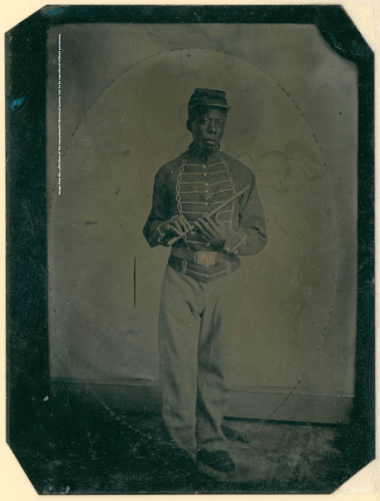 Young boy wearing a jacket with piping braided from shoulders to waist, and piping running from braid to each button on both sides. He wears a belt and a kepi, and holds a fife in both hands.