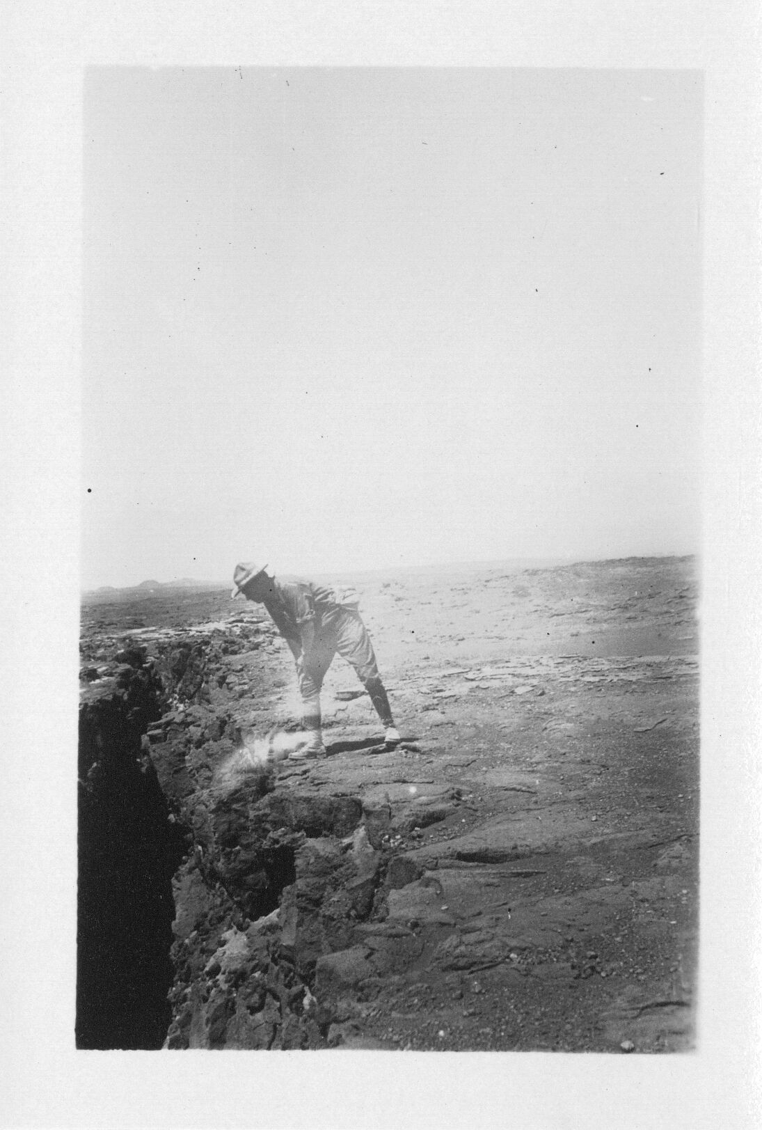 A black and white image of Thomas Boles looking inside a pit crater in the Kaʻu desert. The pit crater is located on the left side of the image. Thomas Boles is in the center of the image standing at the edge of the pit crater. He is wearing a hat, a long sleeve shirt with the sleeves rolled up, jeans, and boots. There is a bag on his back. Boles is in a wide-legged stance facing the camera, his torso is turned to his right leaning towards the pit crater. His hands are resting on his right kneecap, that leg is bent slightly. The lava surrounding the pit crater is smooth pāhoehoe lava.
