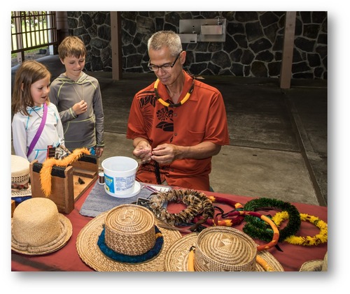 A man wearing a feather necklace sits at a table weaving a feather lei, while children watch. Several woven hats with feather hat bands are placed on the table with feather leis. 