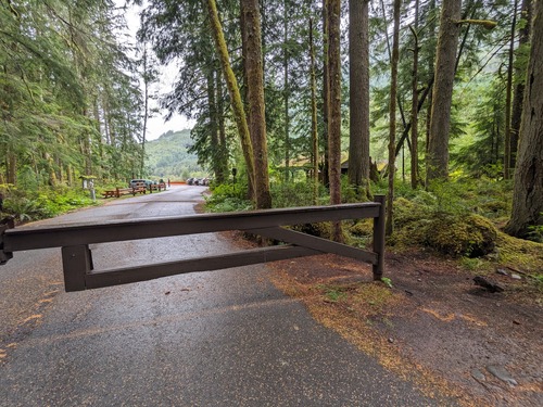 A brown metal gate is closed across a paved road. A narrow dirt path around the gate post allows trail access to pedestrians and cyclists.