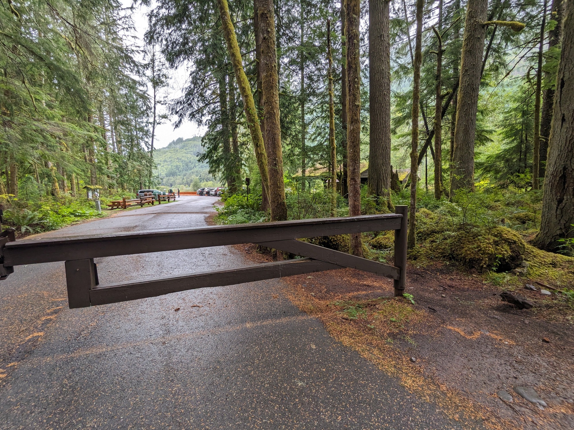 A brown metal gate is closed across a paved road. A narrow dirt path around the gate post allows trail access to pedestrians and cyclists.