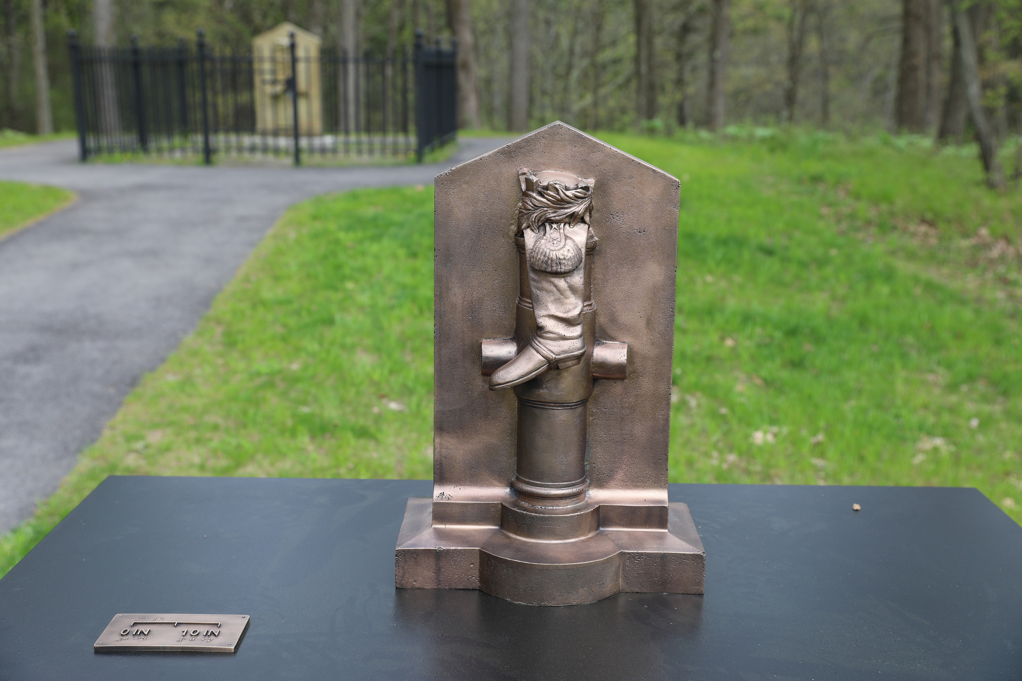 A bronze casting of a monument with a boot laid over a cannon with military epaulettes, and foliage laid on top of the boot. All of these objects are placed in front of rectangular background that comes to a point at the top. This bronze casting sits on top of a black table with a measurement scale and braille plaque next to it.  