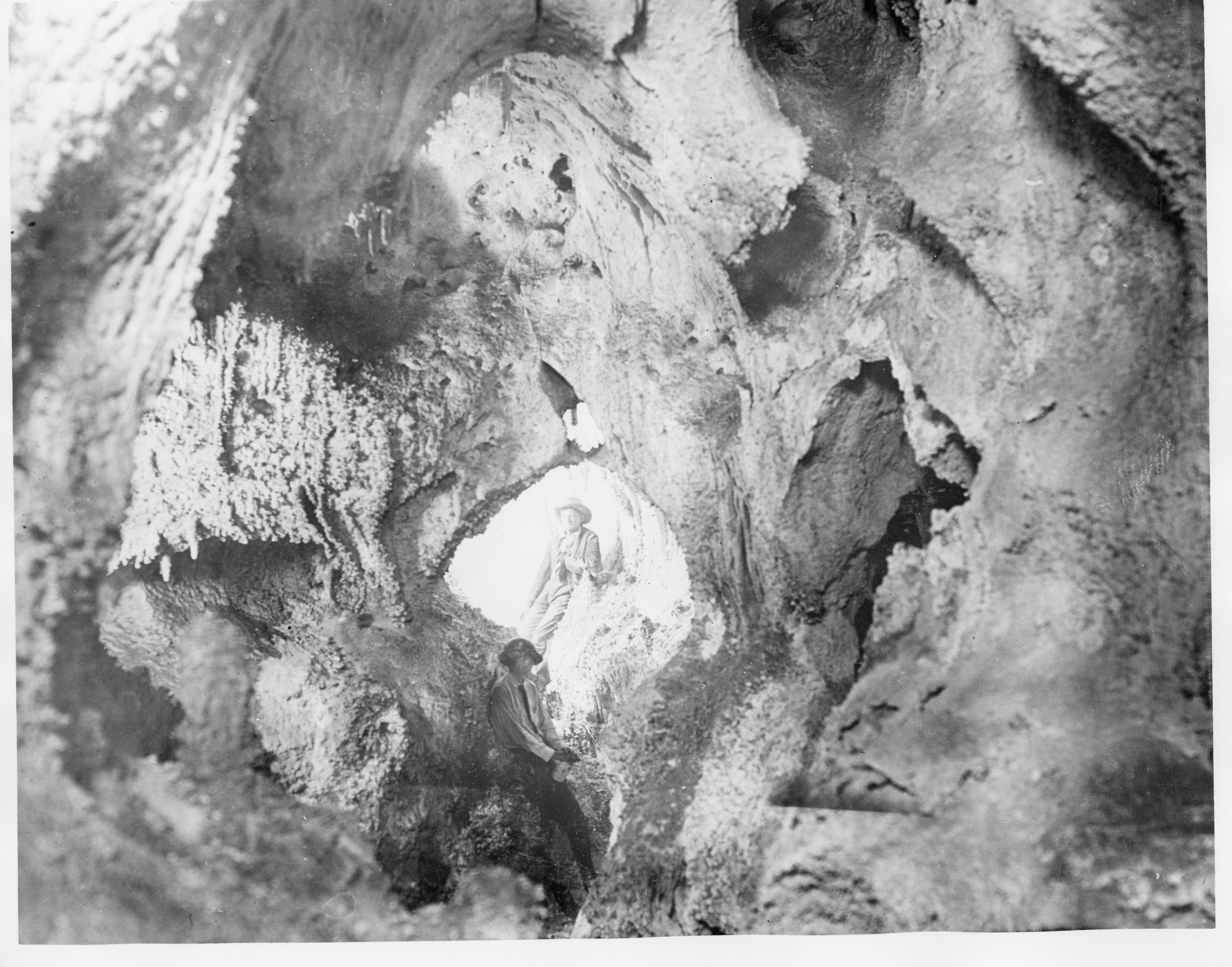 A black and white photograph of two people leaning up against formations to a lighted rock entrance.