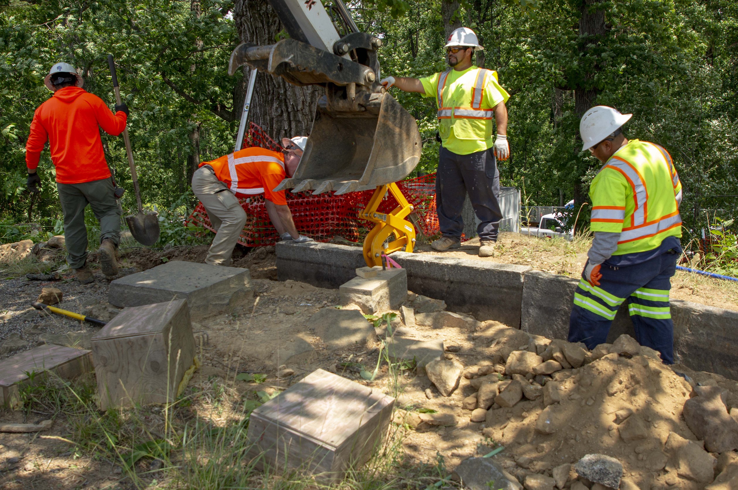 Four contractors, two in green and two in orange, guide the arm of a piece of construction machinery that lowers granite curbing into a trench in front of a tree. 