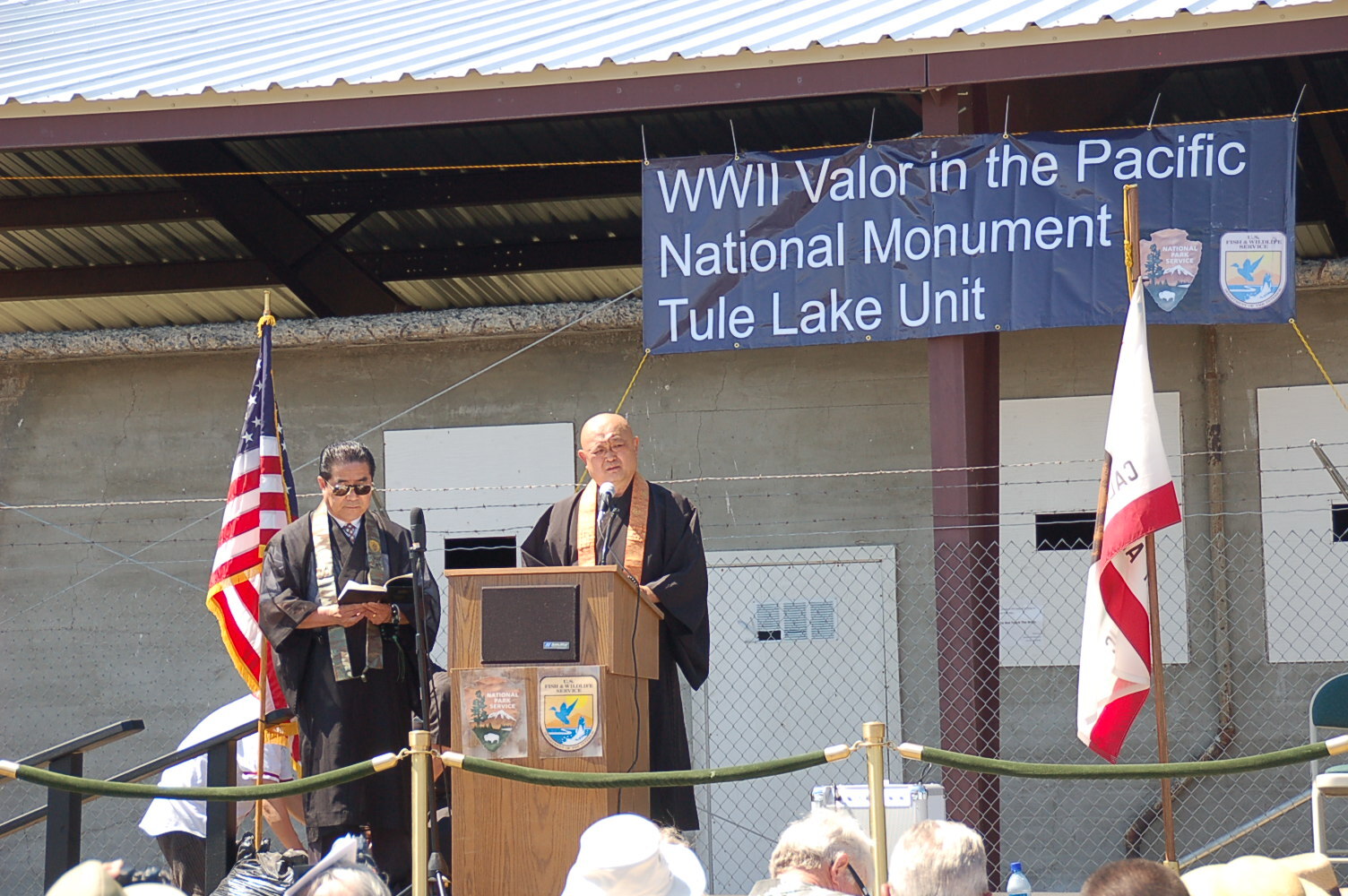 Two men in religious clothing stand in front of a podium by U.S. and California flags. In the background is a building with a sign reading WWII Valor in the Pacific National Monument Tule Lake Unit