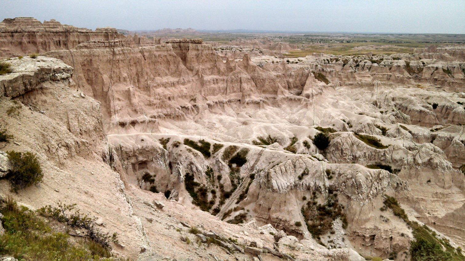 A large ladder climbs up a steep formation, surrounded by more badlands formations