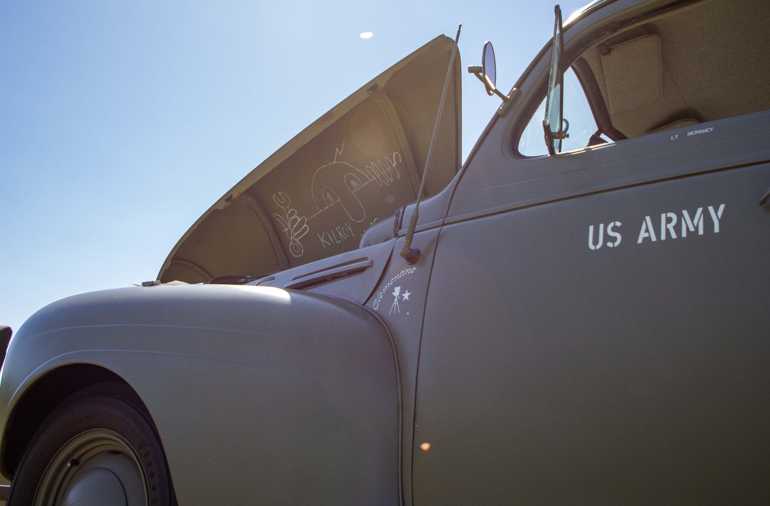 A brown WWII era vehicle with US Army on the side. Under the open hood is a drawing of a figure with text "Kilroy was here"