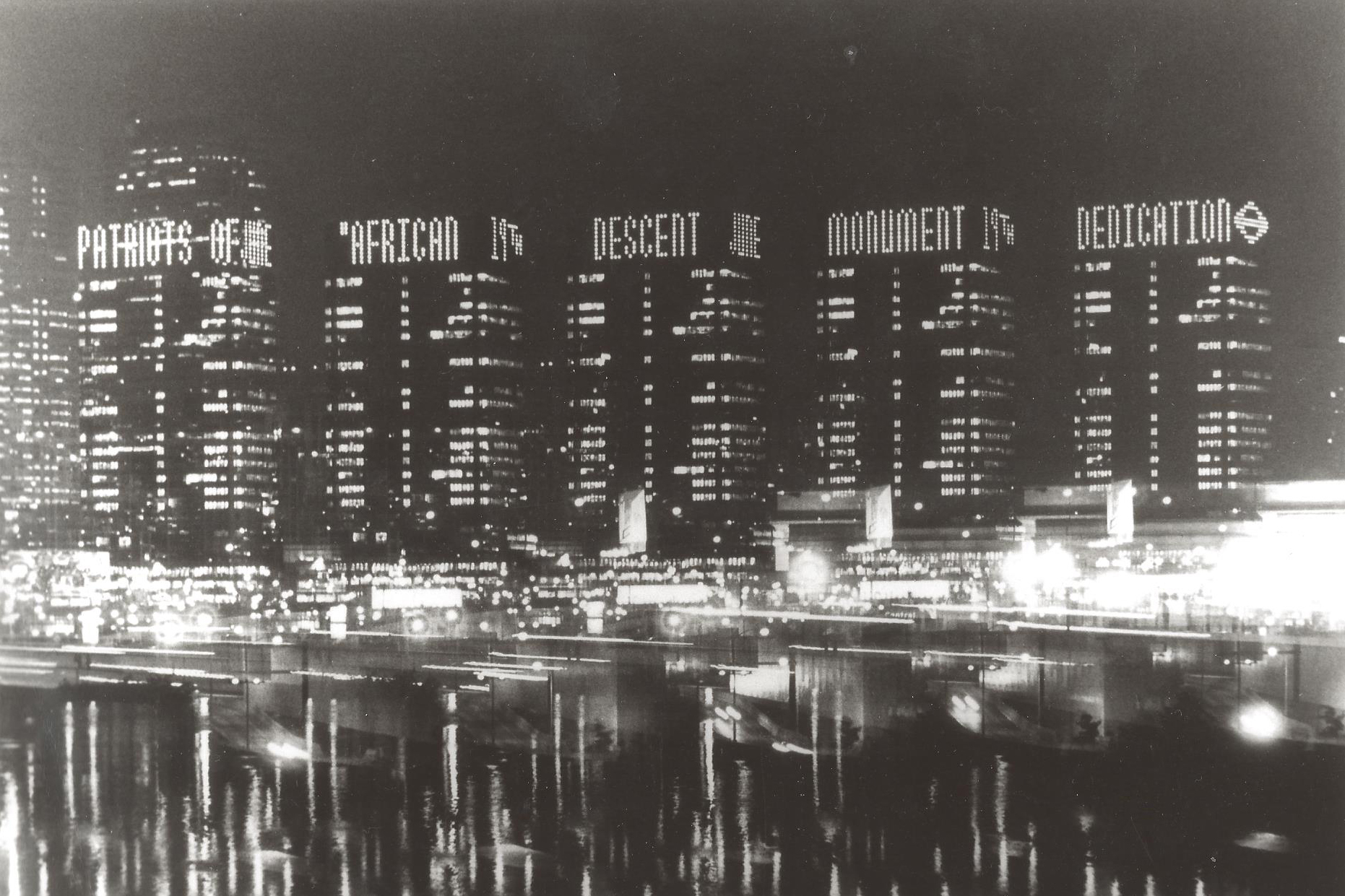 photograph, black and white, nighttime, skyscraper buildings, river with reflected city lights, billboard lighting on buildings spell Patriots of African Descent Monument Dedication, June 19th.