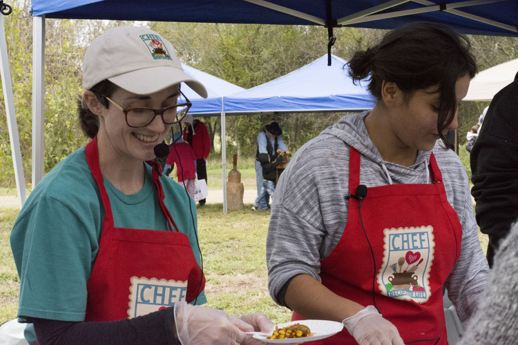 Two women with cooking aprons, one holding a paper plate of a food sample.