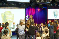 Many children raise their hands as they look toward a park ranger who leads them in a Junior Ranger program.