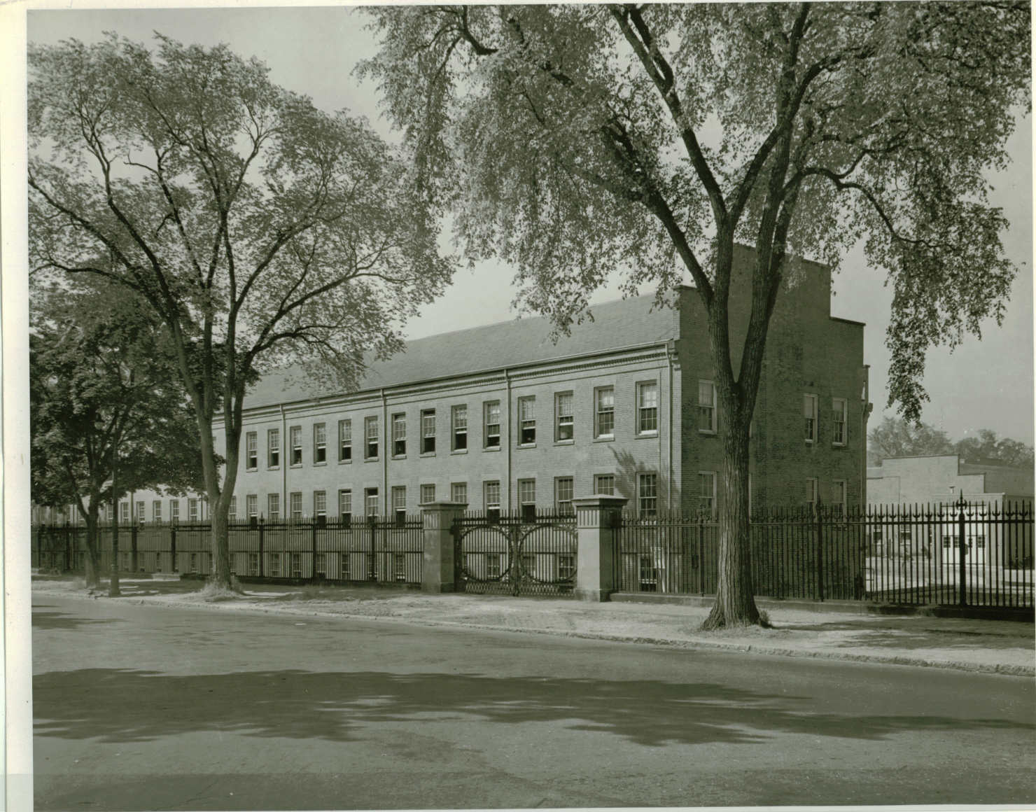 Black and white photo take from across a road that looks at a large brick building behind a metal fence. The fence has a gate with double doors. Trees grow along the roadside.