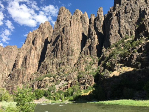 Canyon walls loom above from river level view. Blue sky and clouds appear behind walls.