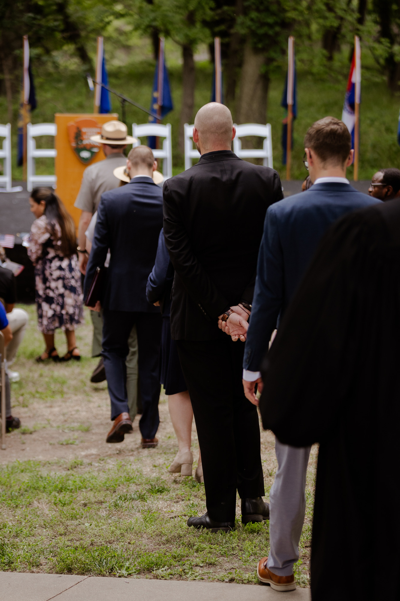 Three individuals wearing suits follow two uniformed park rangers down a center aisle toward an outdoor stage.