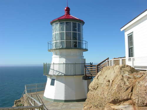 A three-story, sixteen-sided tower with white sides and a red roof. The third level is surrounded by windows. To the left is the Pacific Ocean; on the right is the corner of a white building atop a rock outcrop in the foreground.