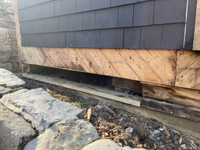 The base of a dark brow stained shingle sided wall with lower shingles stripped away to reveal the wall underneath made of bare boards slanted downward from left to right. Below that, rotten framing boards and beams are visible and are in the process of being replaced. The top of a stone retaining wall is visible crossing the bottom right quarter of the frame from top left to bottom right, parallel to the wall.