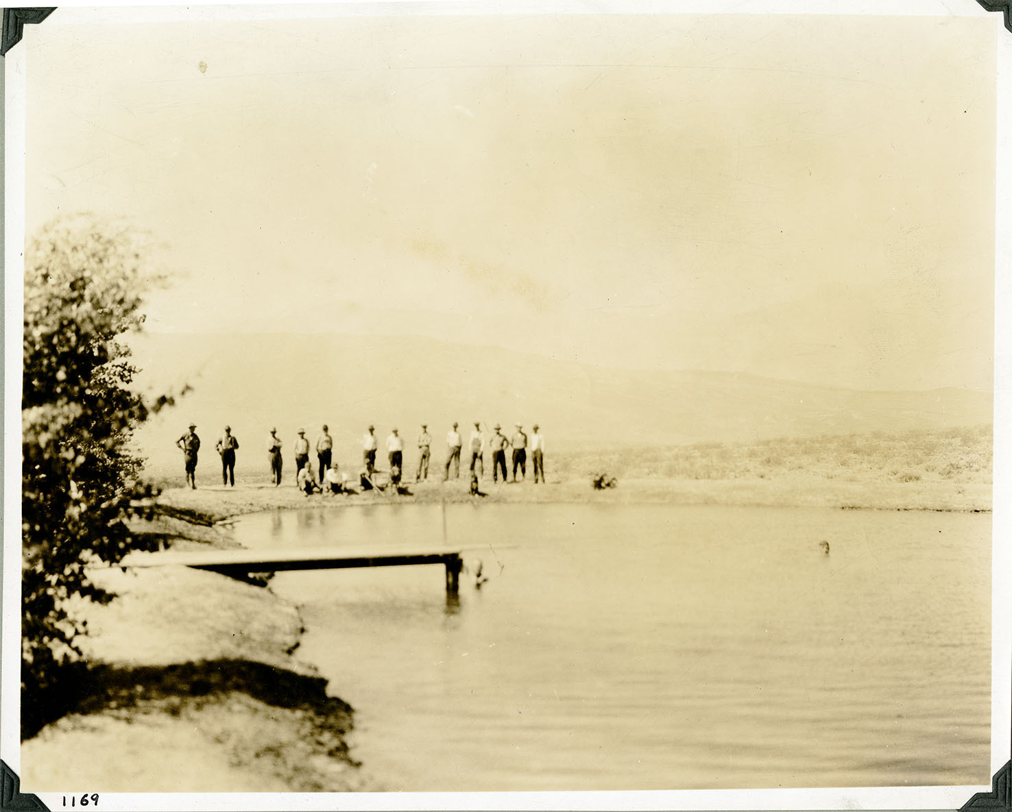 This is an historic black and white photograph from the Scotty's Castle Historic Photograph Collection, Death Valley National Park of at least sixteen men standing and sitting on dirt dam behind reservoir. Wood pier over reservoir to left. Two men in the water. Two dogs with the men. Vegetation on left side of earthen berm. Number in black ink in lower left corner.