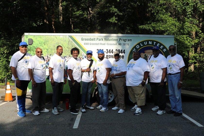 A group of ten men wearing white t-shirts stand in front of a white trailer that reads "Greenbelt Park Volunteer Program." The men are of varying builds and ages, and some wear hats. The background features green trees and a paved area with an orange traffic cone.