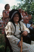 An old woman wearing a black bonnet and long sleeved dress, sitting near other people at an outdoor event.