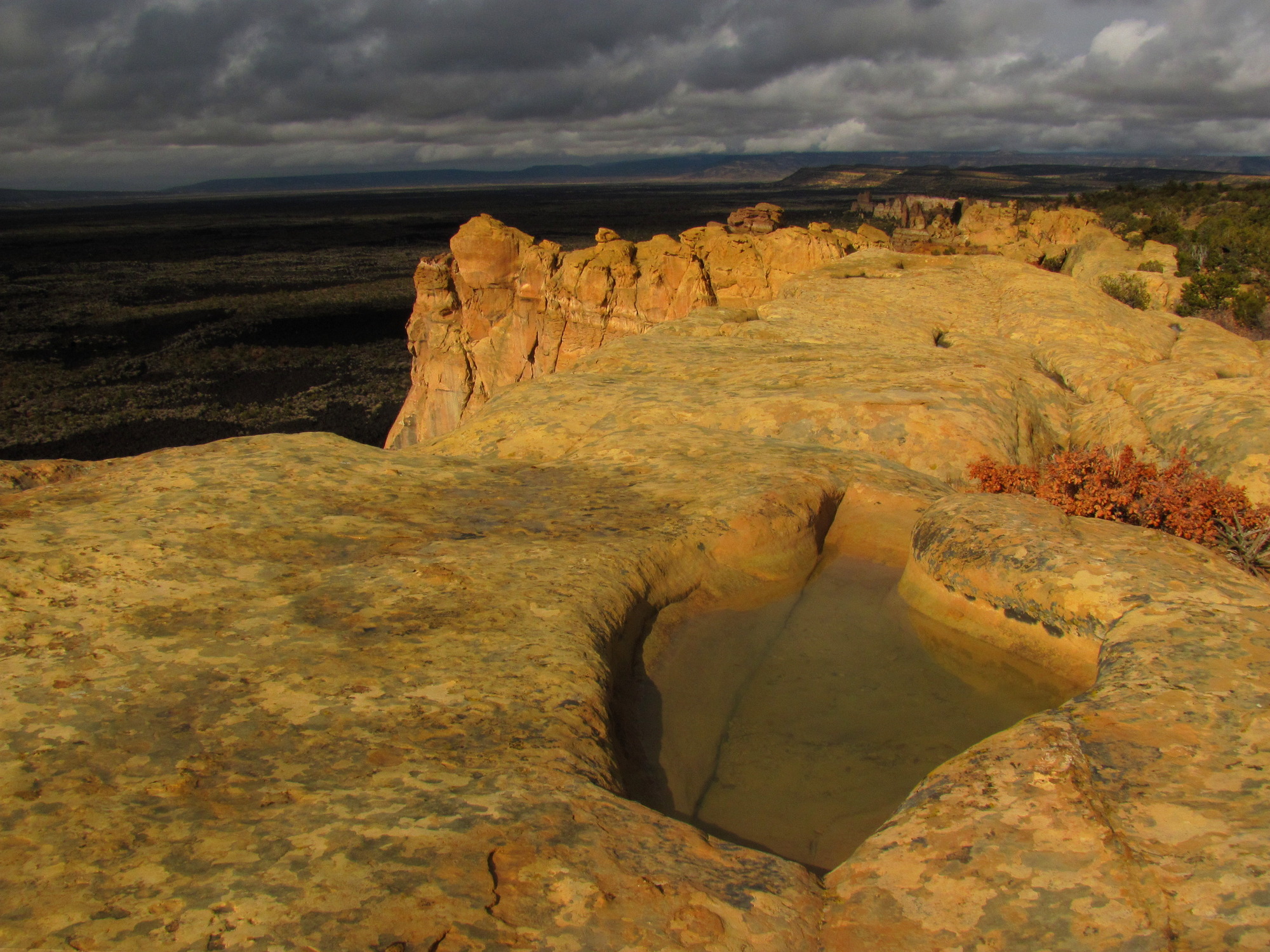 Dark clouds cast the shadow of incoming rain across yellow sandstone.  An oval-shaped depression in the sandstone stands half full with water.