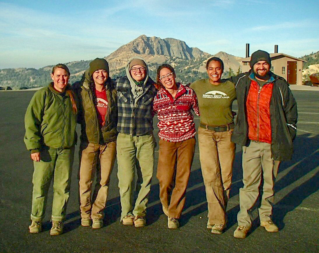A group of six young men and women pose for a photo in a parking area, backed by a large gray mountain. 