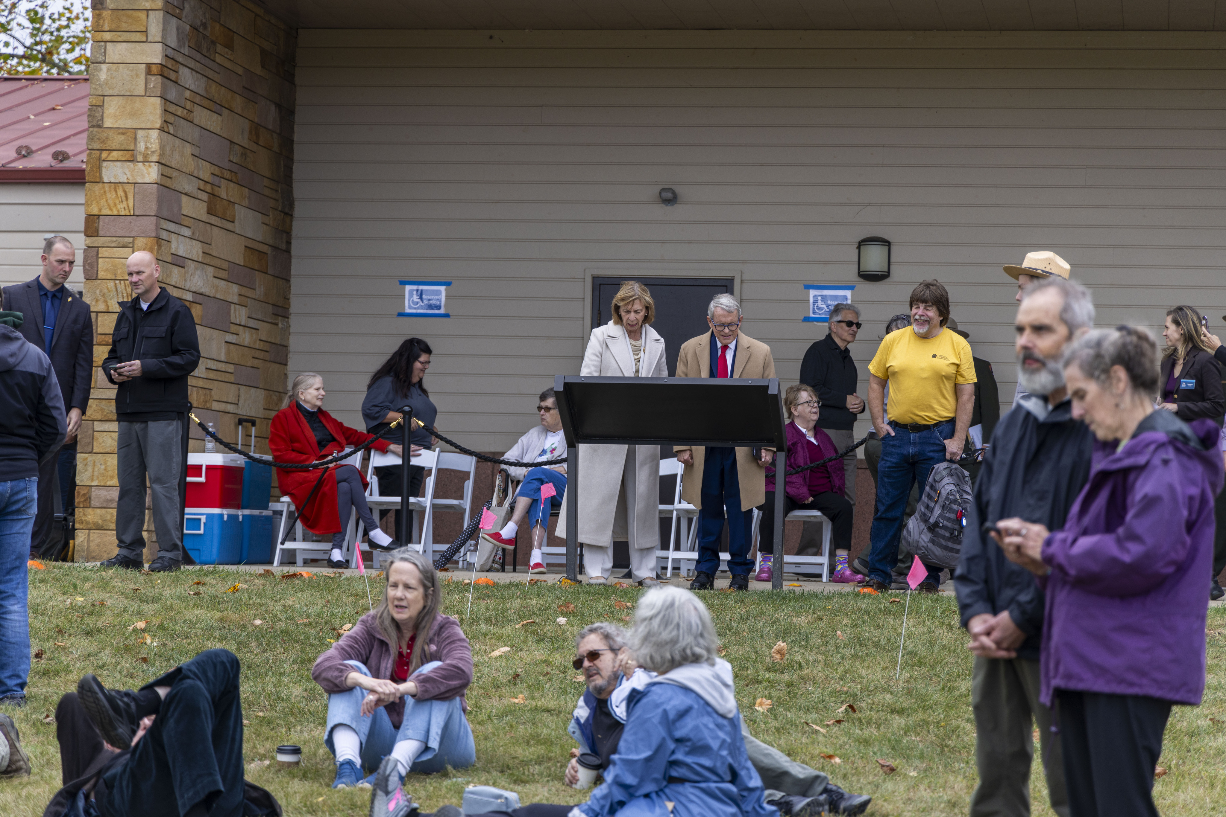 In the middle of the photo, a man wearing a brown long coat and a woman wearing a white long coat standing in front of a black panel reading its contents. Several people stand and sit in various spots of the photo.