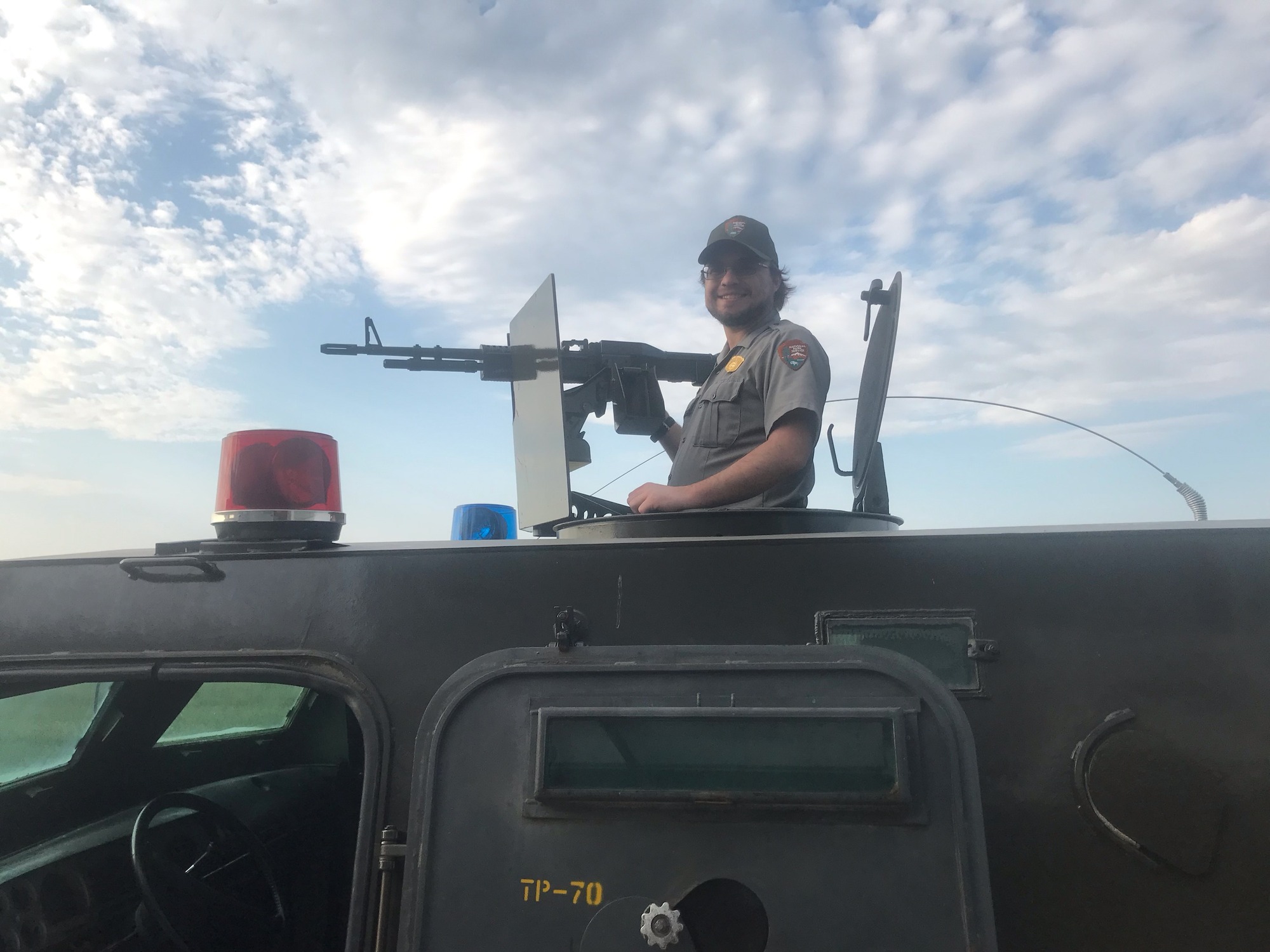 Man in a gray shirt and green cap standing in gun turret on a green vehicle with a machine gun
