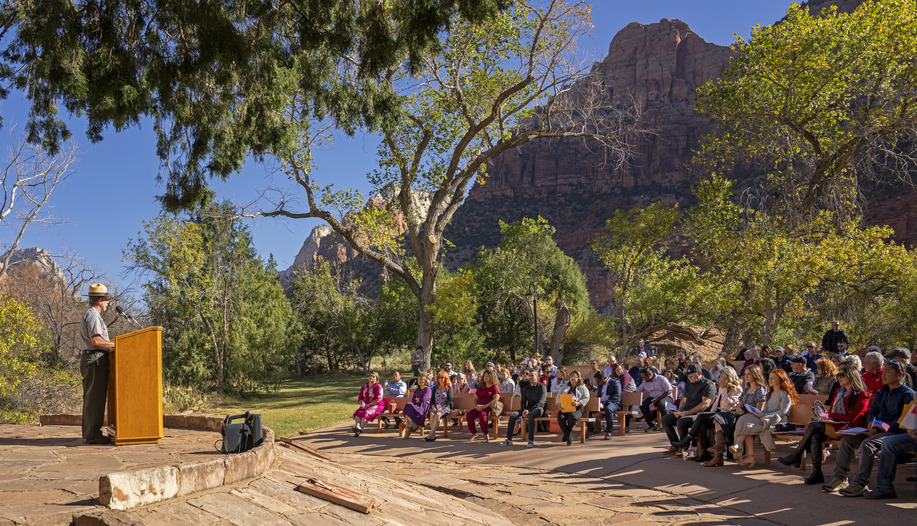 A man in a park ranger uniform and hat stands at a wooden podium, facing an audience of seated people surrounded by green trees. 