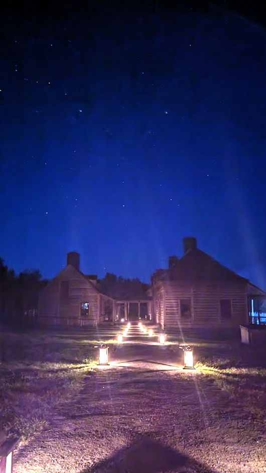Candlelit lanterns line a walking path to a group of historic buildings under a starlit night sky.