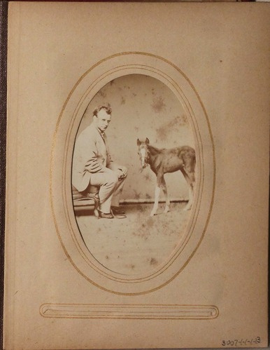 Black and white photograph of man seated looking at the viewer, across from him is a small horse.