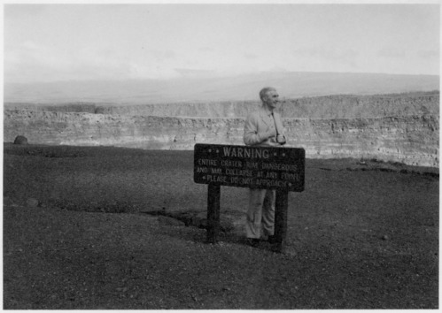 Black and white. Man in light colored suit standing behind a wooden sign with crater in the background