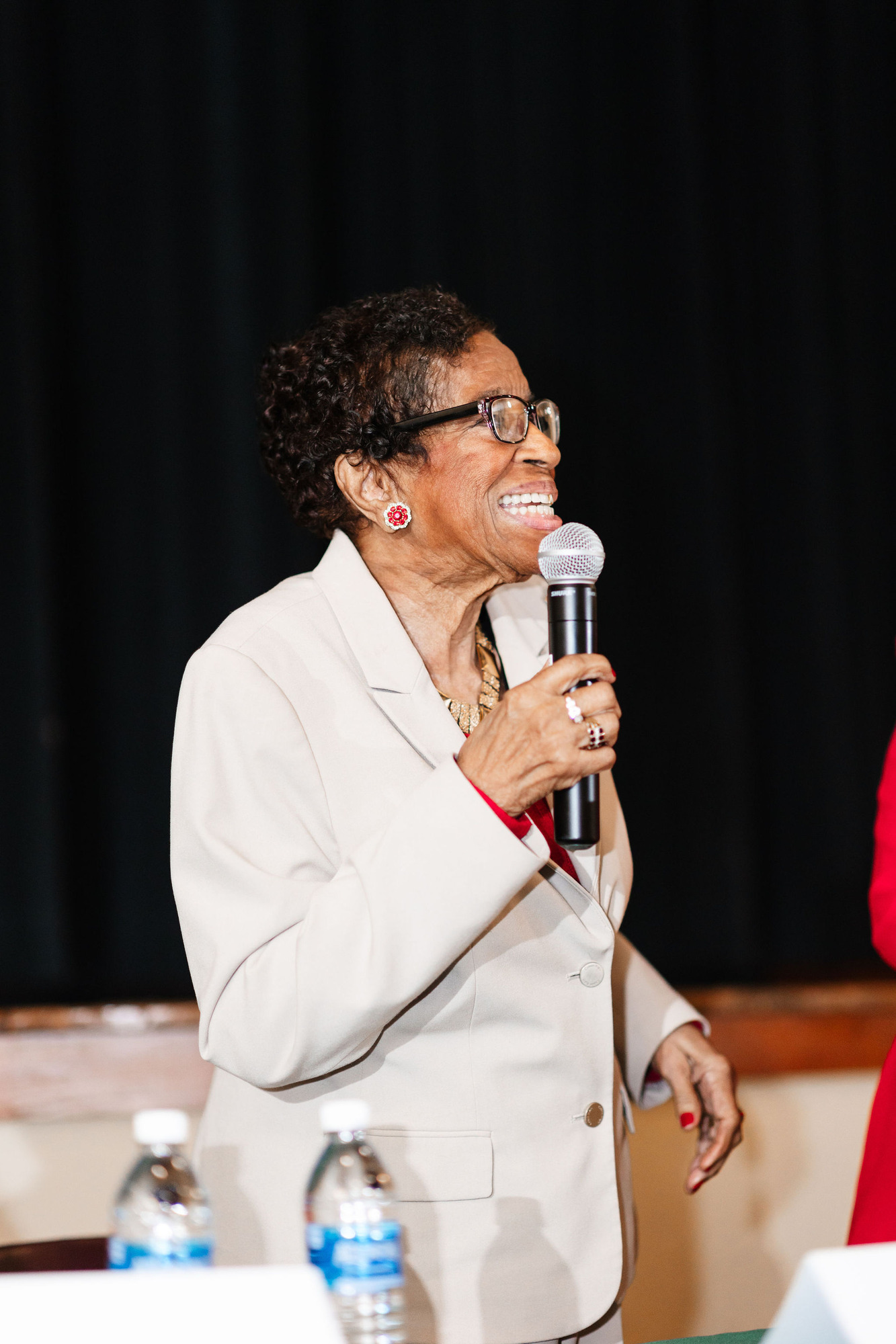 An older african american woman in a white suit stands in front of a black curtain while speaking into a microphone
