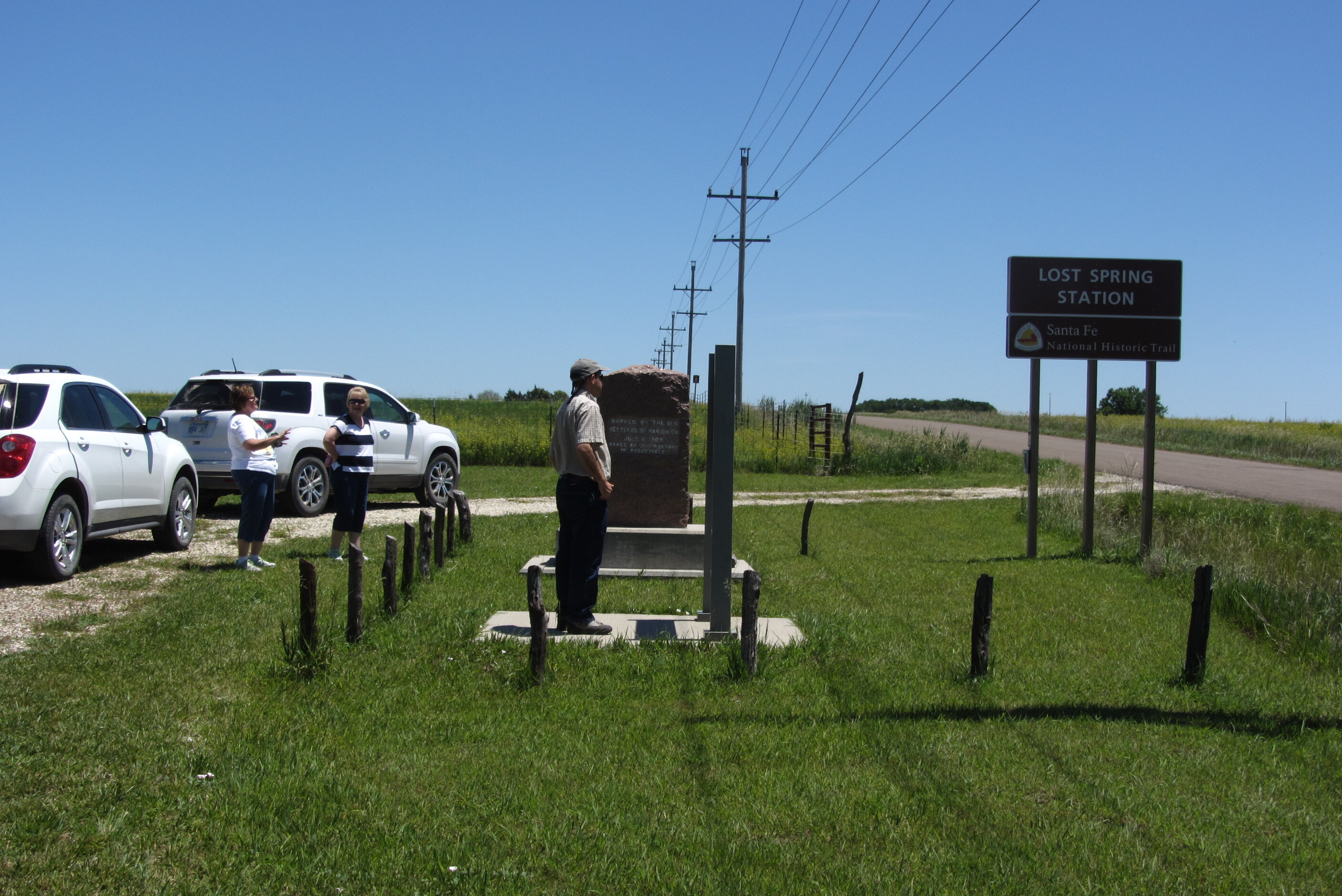 A group of people next to an informational sign titled "Lost Spring Station" with text and illustrations on it beside a road.