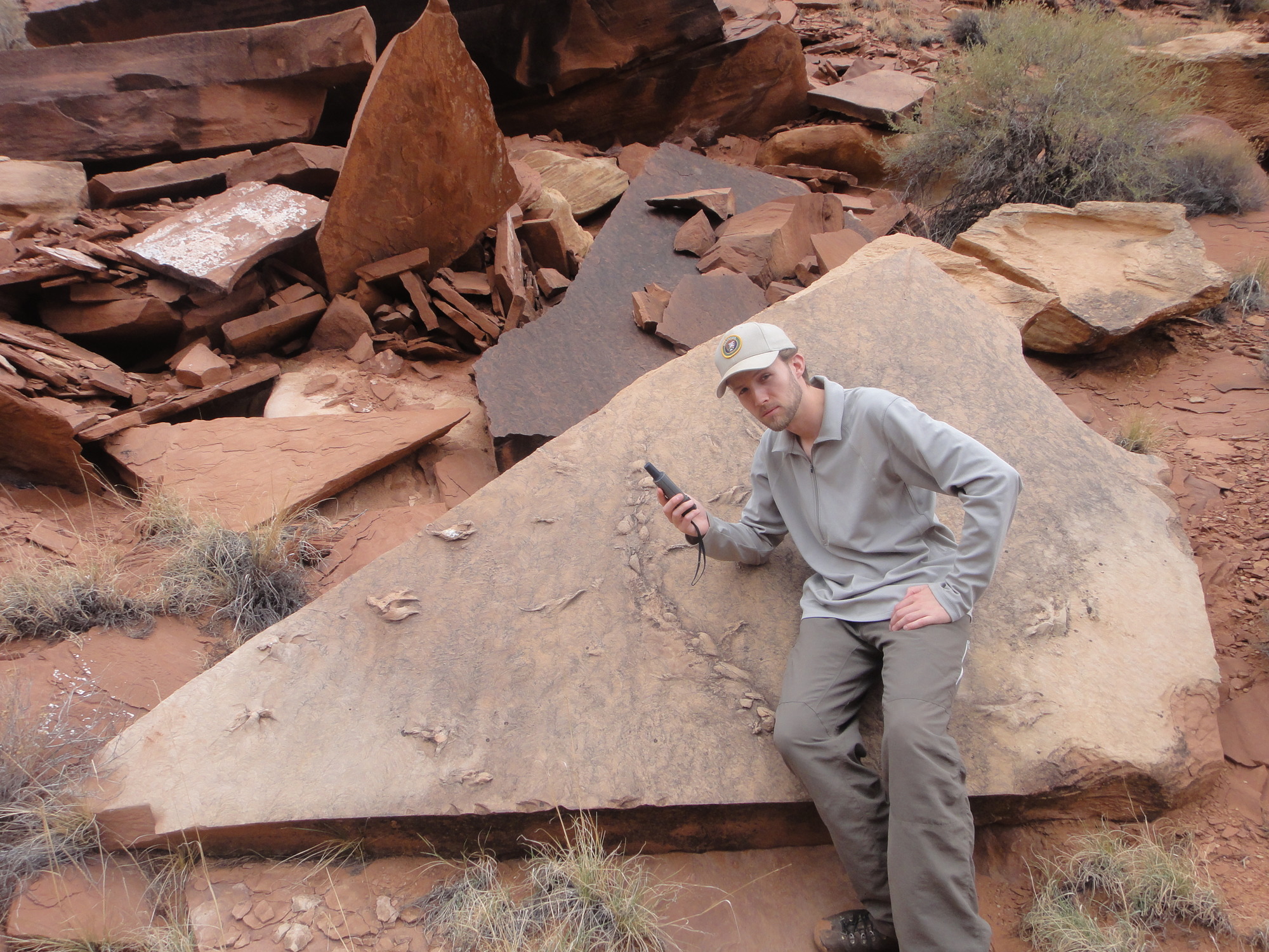 a young man holds a GPS instrument while leaning up against a wedge shaped rock within a pile of rocks. 