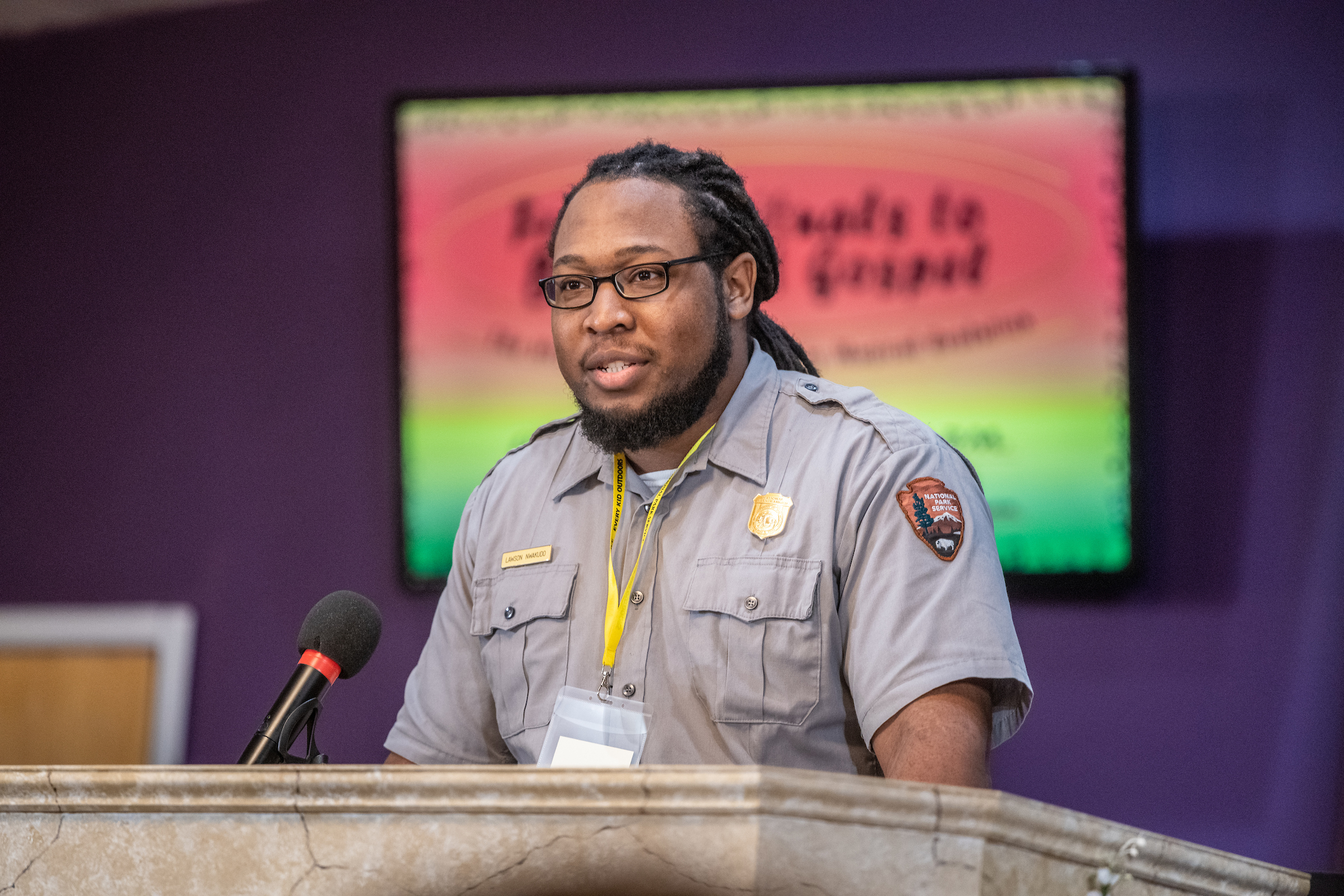 Close up of Ranger Lawson Nwakudo giving an interpretive talk. The red, yellow, and green event graphics are visible in the background.
