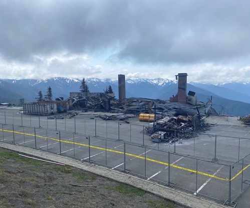 A chain link fence encloses the burned wreckage of a building, two chimneys still standing amid the rubble. Snow-capped mountains stretch into the distance.