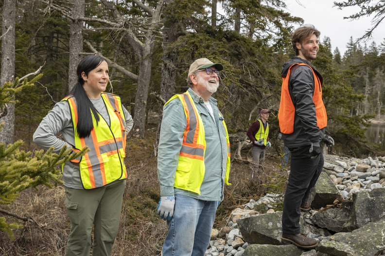 Volunteers in high-visibility vests engaged in a cleanup effort in Acadia National Park, with a focus on three individuals in the foreground smiling and looking off to the sides in a forested environment strewn with rocks and boulders.