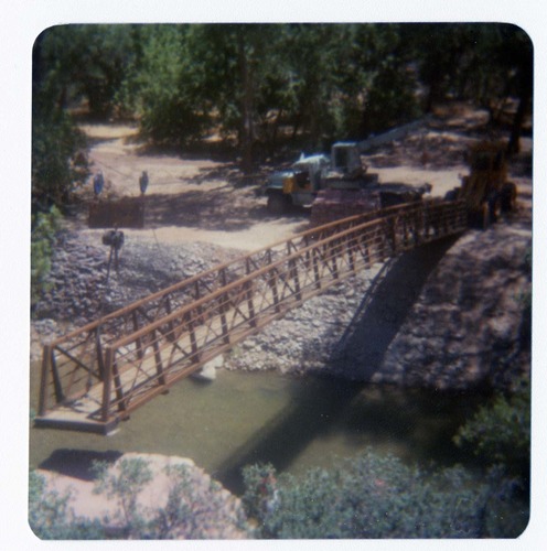 New Grotto footbridge being lowered into place by pulley system, crane and backhoe anchoring one end of bridge. Note riverbank revetments.