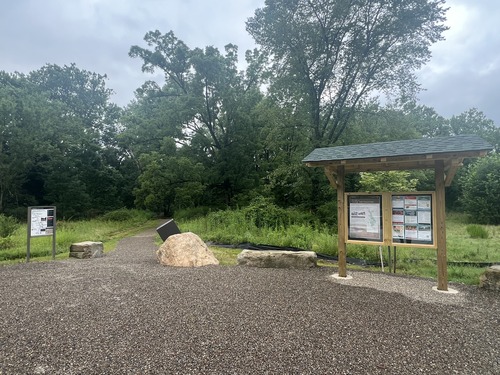 A map panel is left of an unpaved path into trees. Right is a kiosk with a welcome panel and flyers.