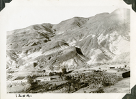 This is an historic black and white photograph from the Scotty's Castle Historic Photograph Collection, Death Valley National Park of a large l-shaped building with a flat roof setting along the length of a wash at base of barren, steep desert mountains. Another building at right edge is a barn. Dense vegetation between the two structures. Plane prop on right edge.