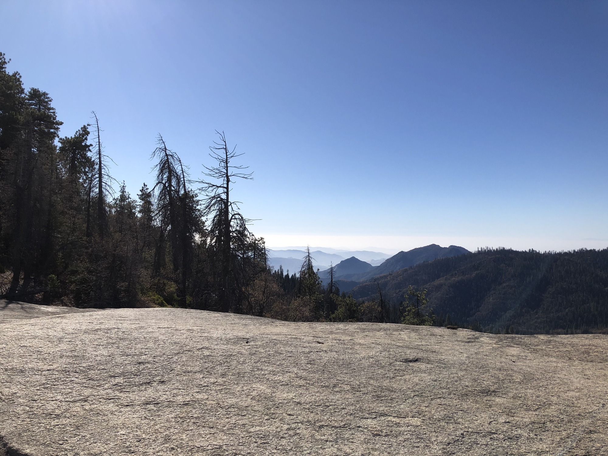 A flat expanse of granite with trees and hazy views of faraway hills