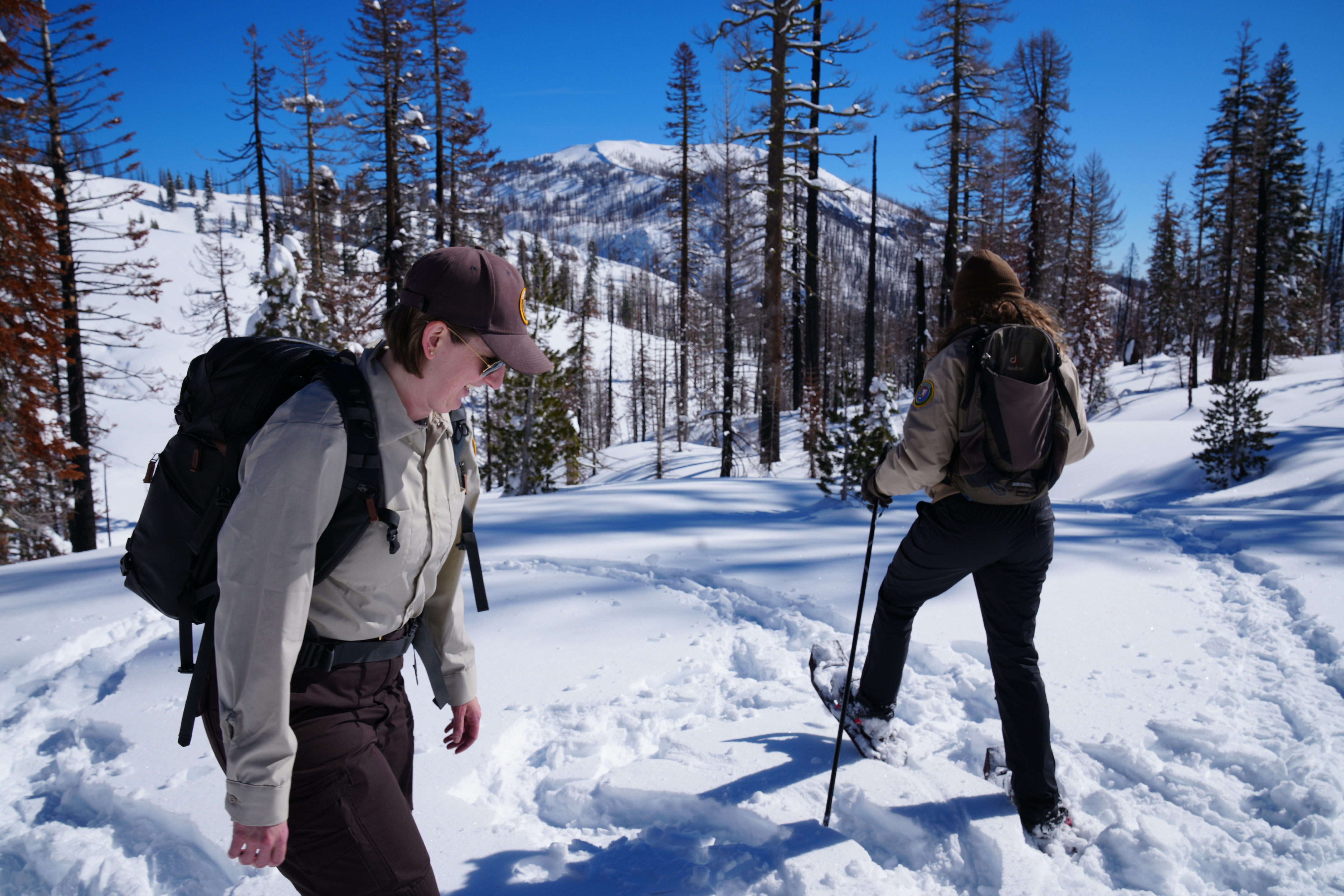 Two volunteers walk through the snow on snowshoes. A mountain can be seen in the background. The volunteer in the front is looking down smiling, while the volunteer in the back has her back to the camera and is creating a trail of snowshoe prints.